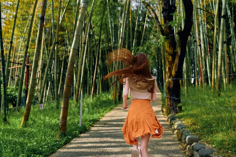Woman running through a bamboo forest on a gravel path, wearing an orange skirt with red hair flowing, back view in nature capturing summer outdoor motion and joyful movement.