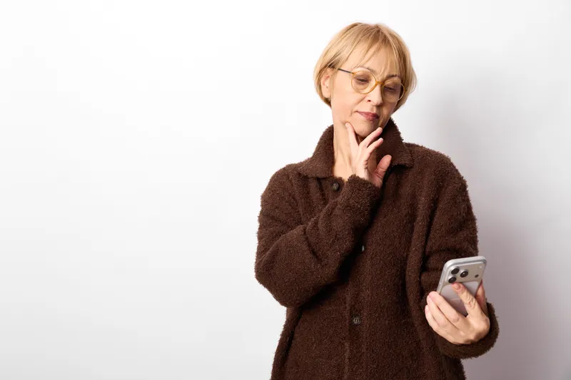 Woman holding smartphone and touching her face, thinking on a bright white background. Close portrait with brown coat, calm mood, indoor lifestyle carefully