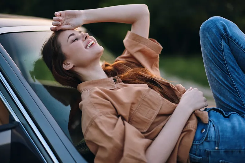 Young woman enjoying a sunny day, dressed in a casual brown shirt and blue jeans, expressing joy while resting on a car hood against a natural green background.