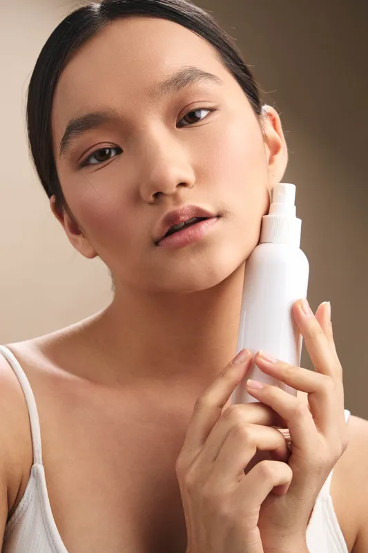 Asian woman holds a white bottle close to her face, showcasing skincare product. She gazes softly at the camera, conveying calm confidence and self care in a minimal studio setting.