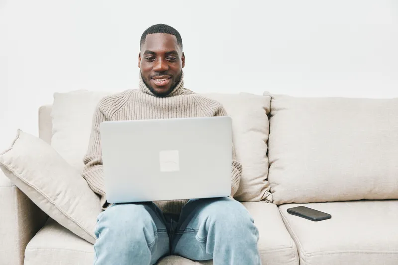 Smiling Black man working on a laptop at home in a cozy sweater, reflecting productivity and comfort, perfect for remote work or online learning
