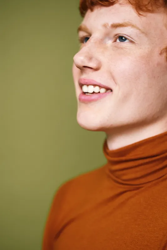 Portrait of smiling young man with red hair wearing brown turtleneck against green background. Closeup of happy male face with clear skin and expressive eyes looking up and right.
