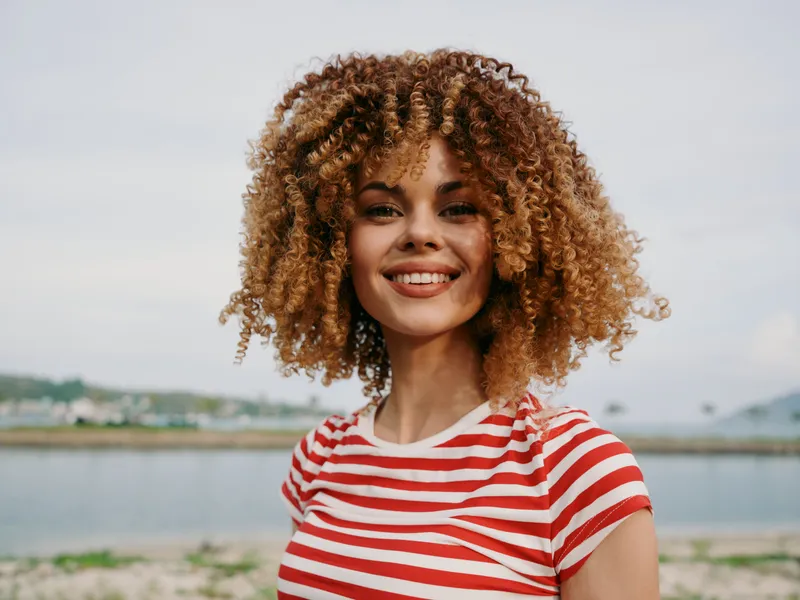 Portrait of a cheerful young woman with curly hair wearing a red and white striped shirt, smiling at the camera on a sunny seaside setting.