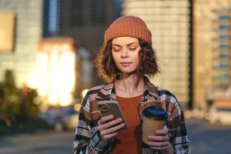 woman smartphone coffee urban beanie plaid goldenhour candid: Young woman in plaid shirt and beanie checks her phone holding takeaway coffee in warm golden hour glow, authenticity, candid lifestyle