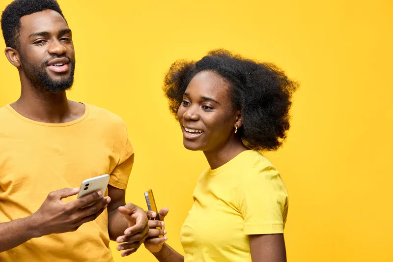 Happy young couple enjoying a playful moment with smartphones against a vibrant yellow background, showcasing love, connection, and joy in casual attire