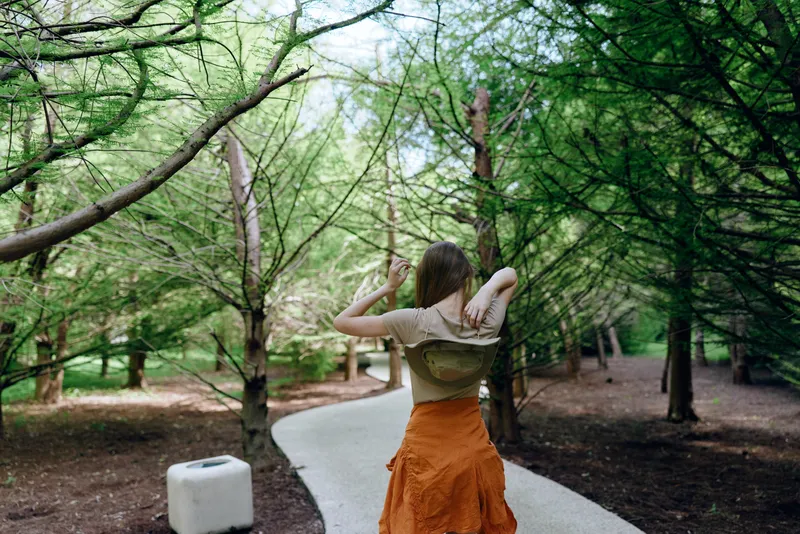 Woman walking on a forest path with backpack among green trees, trail winding ahead, casual outfit and orange skirt, adjusting hair on a summer outdoor hike in peaceful nature.