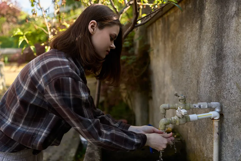 Smiling woman in a plaid shirt washes her hands at an outdoor faucet, sunlit wall behind. Closeup scene emphasizes daily care, cleanliness, and simple wellness