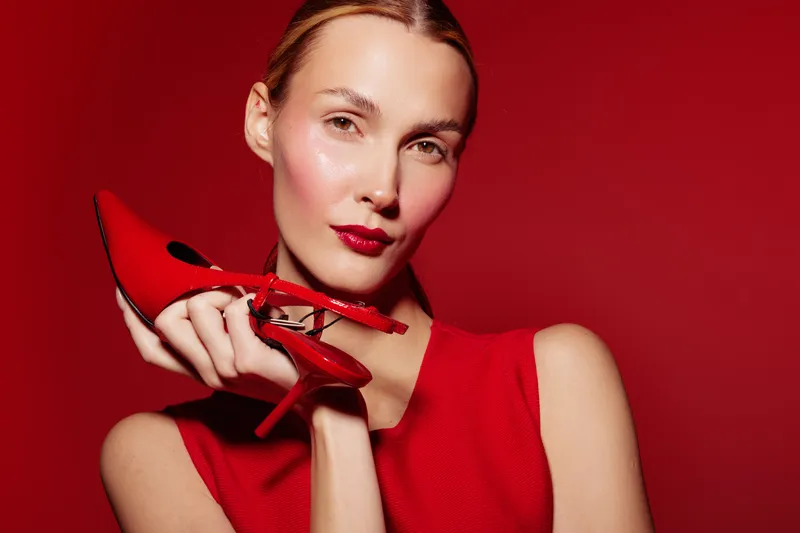 Stylish woman in red dress holds red high heel shoe near face on red background. Concept of fashion, elegance, beauty, and glamour with vibrant color contrast and confident pose.