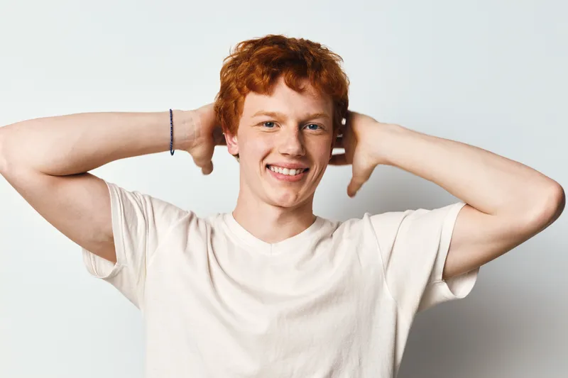 Young man with red hair and freckles smiling with hands behind head in casual white t shirt against white background. Relaxed male posing for portrait with friendly expression.
