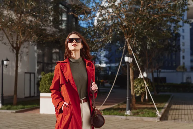 Stylish woman outdoors wearing red coat and sunglasses walking confidently in urban setting with autumn trees and modern buildings in background
