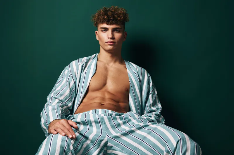 Young man with curly hair wearing striped pajamas sitting against dark green background with serious expression and relaxed posture in studio portrait.