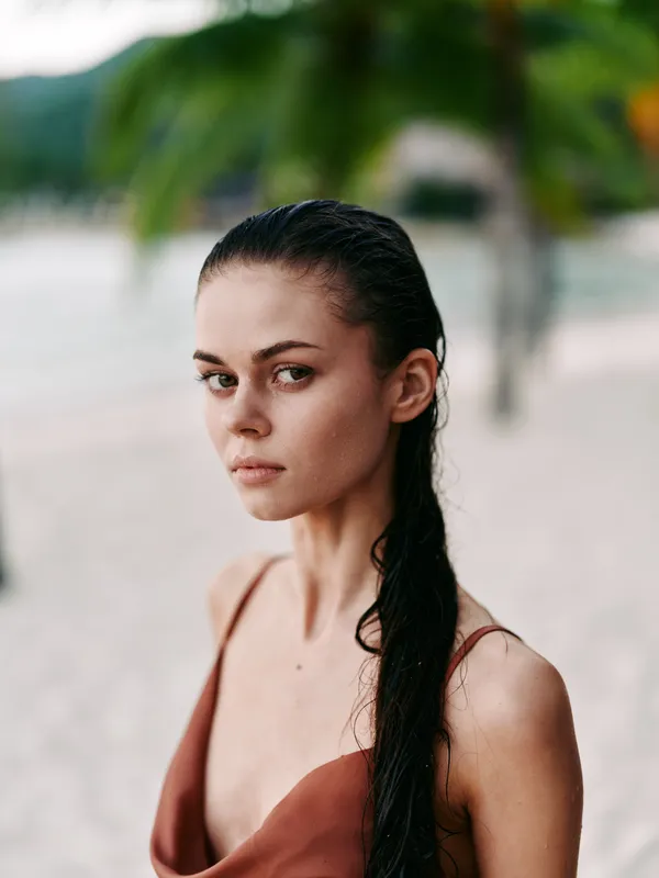 Portrait of a serious woman with wet hair outdoors, wearing a brown top, with a blurred beach background and soft natural light.