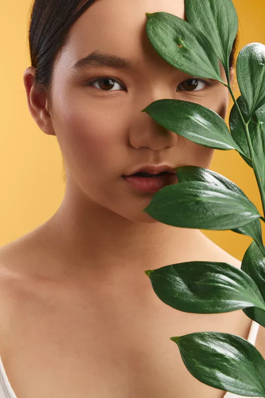 asian woman with smooth skin gazes softly at the camera as leafy greens frame her face, a warm yellow backdrop creates a calm, natural portrait emphasis.