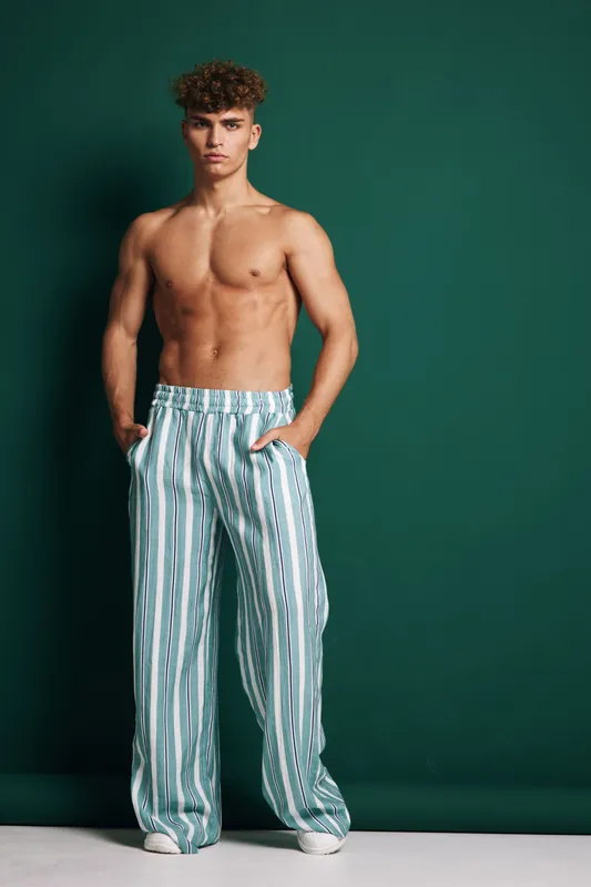 Young man with curly hair posing shirtless in striped wide pants against green background, standing with hands in pockets and serious expression in studio setting.