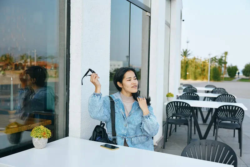 Sunglasses in hand, stylish woman relaxes at an outdoor cafe beside a glass wall, capturing a casual urban moment with reflections, potted plants, and empty chairs under soft daylight