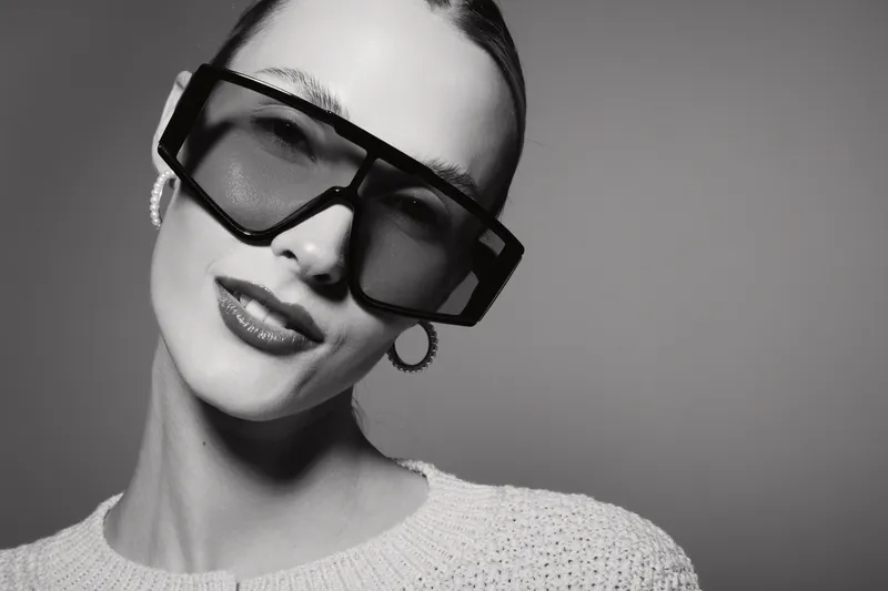Close-up portrait of a young Caucasian woman with fair skin wearing oversized sunglasses and hoop earrings on a neutral background.