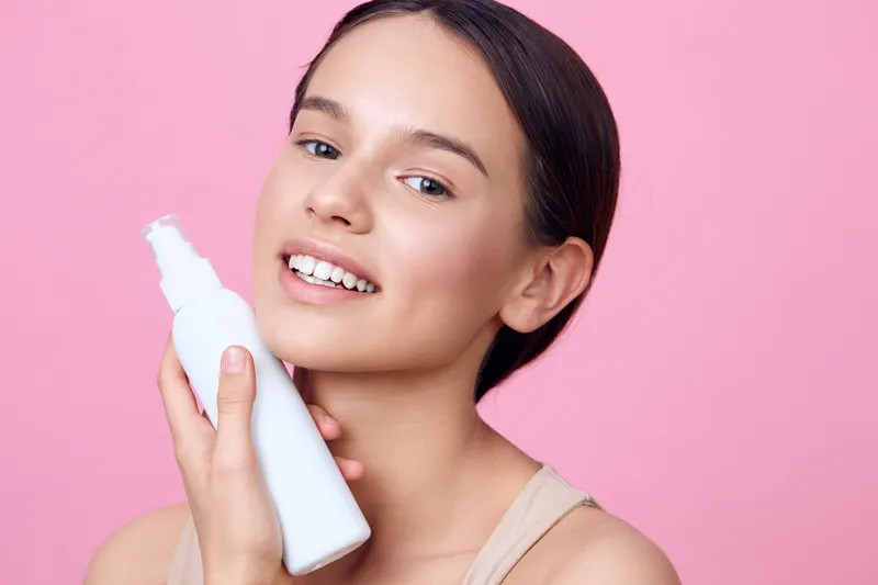 Young woman with smooth skin holding a moisturizing spray on a pink background, representing beauty and skincare concepts with freshness and confidence