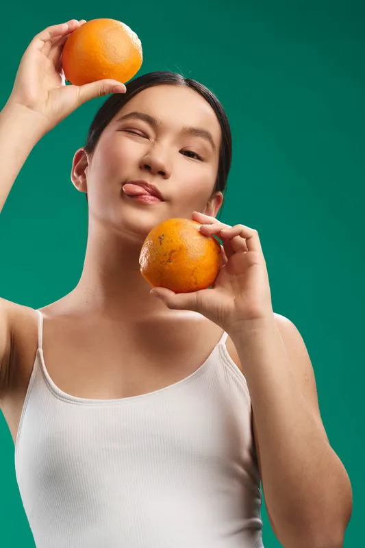 Asian woman is posed in a vibrant studio, holding fresh oranges near her face. She wears a light top and offers a playful, fresh mood with a bright backdrop.