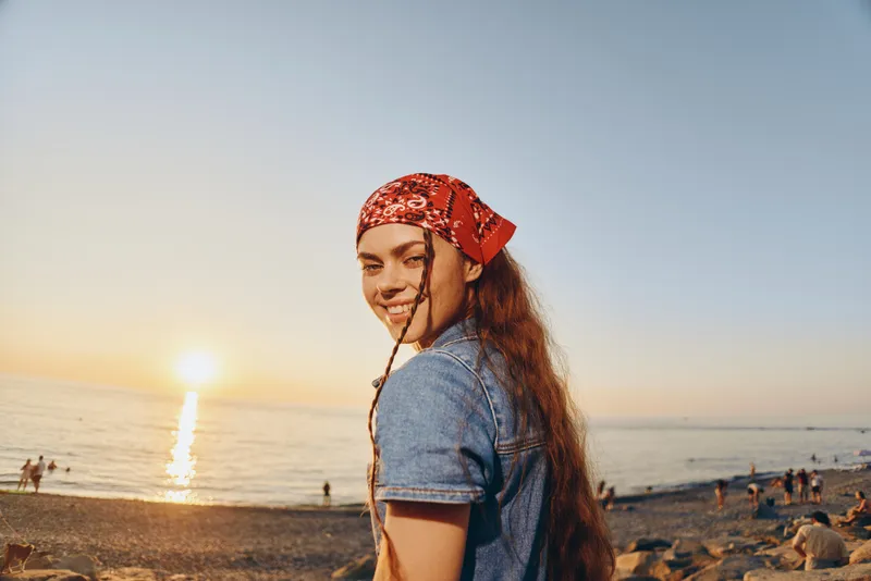 lifestyle woman in modern boho-western denim street style smiling outdoors at sunset with red bandana and natural beach background, film color tone