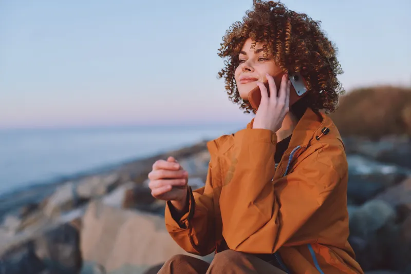 Curly haired woman lifestyle in orange jacket sits on rocky coast, holding a phone to her ear by the sea at sunset, relaxed outdoors communication and calm wellbeing
