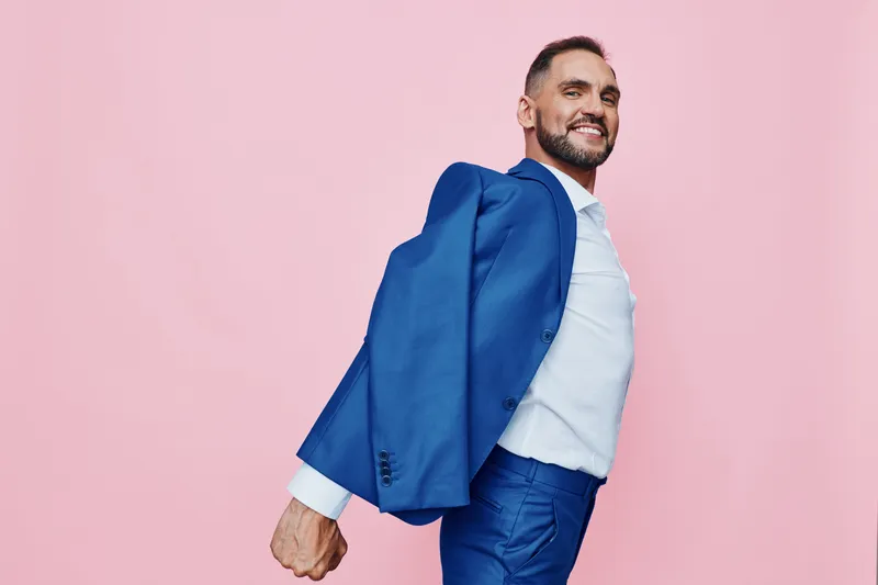 A confident business professional man in a blue suit stands against a solid pink background, exuding energy, optimism, and determination.