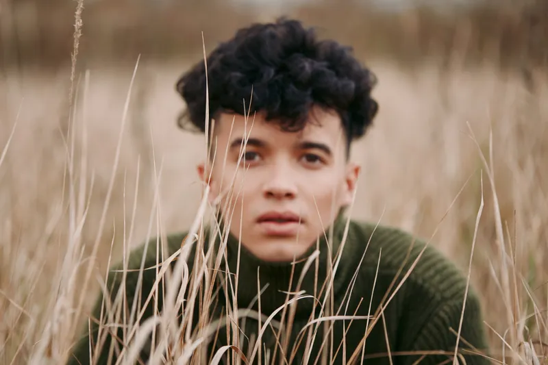Candid outdoor portrait of a young man crouching in tall dry grass, neutral earthy tones and textured sweater emphasizing authenticity and quiet trustworthiness in a natural lifestyle scene.