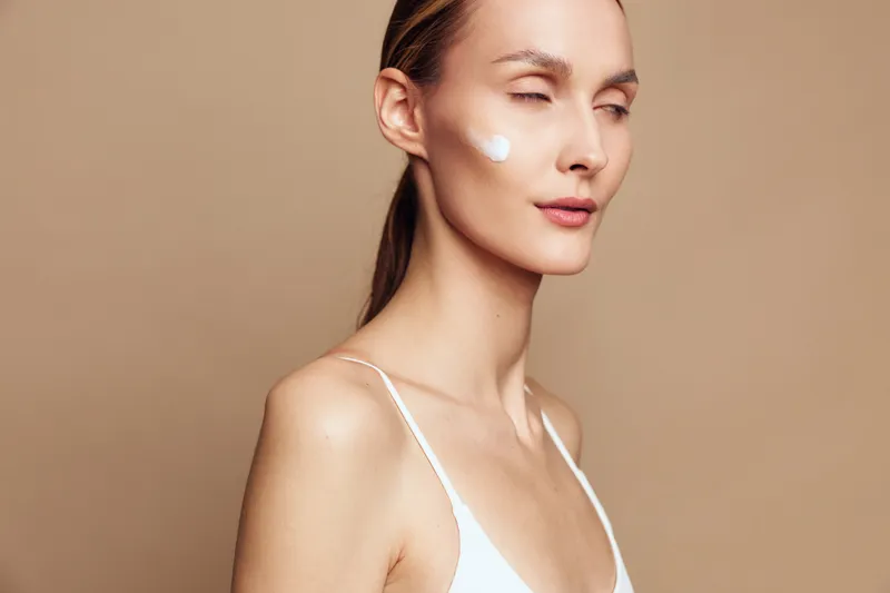 Young woman with long blonde hair applies white face cream on her cheek against a beige background, expressing calmness and skincare routine in soft natural light.