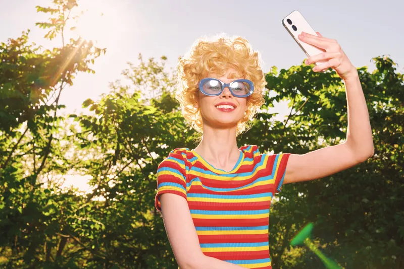 Smiling blonde woman taking a selfie outdoors in a colorful striped shirt. Retro film color mood, sunny setting, playful lifestyle and self expression.