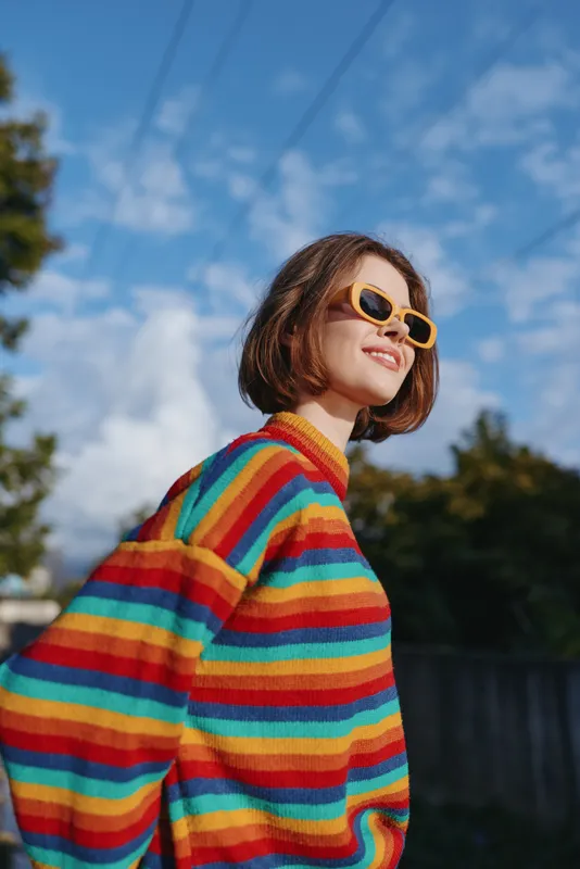 Woman in a rainbow sweater wearing yellow sunglasses, smiling in an outdoor portrait. Casual fashion style, short hair, sunny blue sky background and relaxed confident expression.