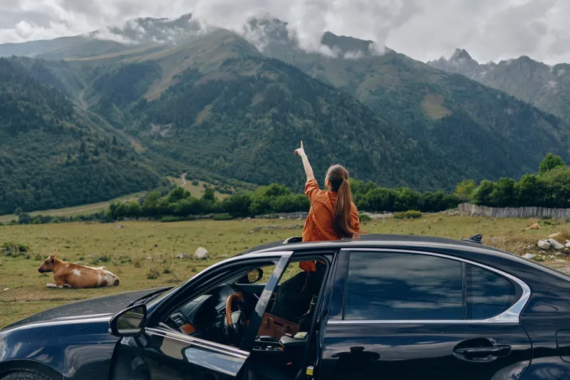 Adventure traveler in a moving car raises an arm through the sunroof as mountains rise over the countryside, capturing a moment of freedom and natural joy