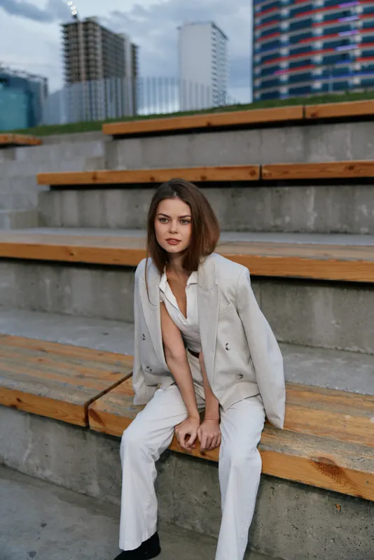 Woman in business casual suit sitting thoughtfully on urban steps with modern city buildings background, showcasing confidence and professionalism
