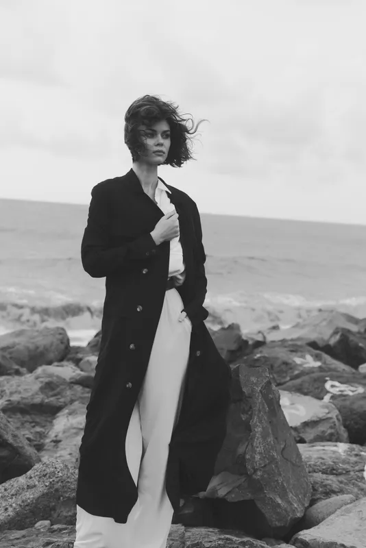 Black and white coastal scene, a woman in a long dark coat stands on rocks by the sea, wind tousling hair, confident pose, minimalist fashion mood at the shore
