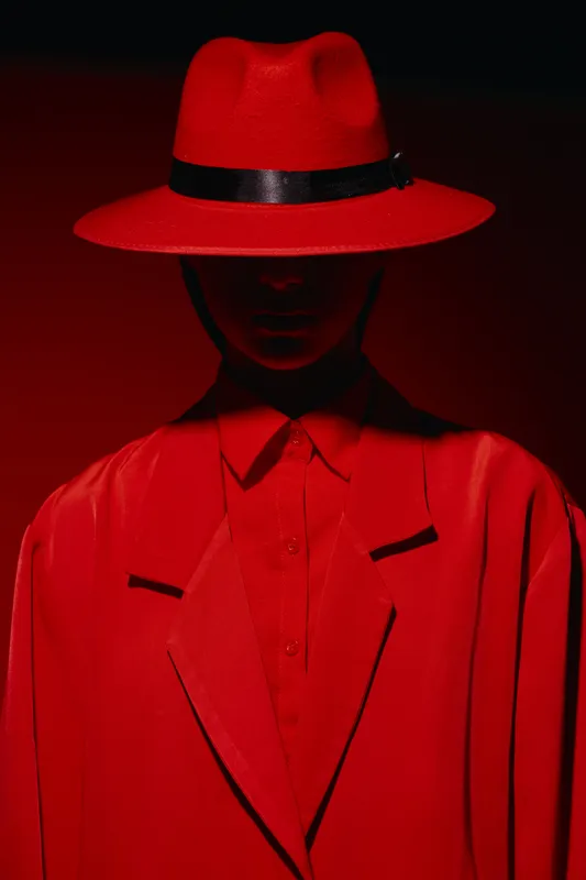 Fashion model dressed in a red hat and coat poses sharply with shadow covering facial details. Image conveys boldness and mystery in a dramatic creative style.