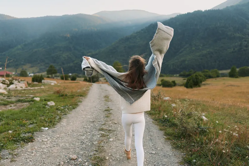 A woman on a rural path raises her arms in joy, embracing freedom and nature. Open landscape, open road, and fresh air capture a carefree lifestyle moment outdoors.