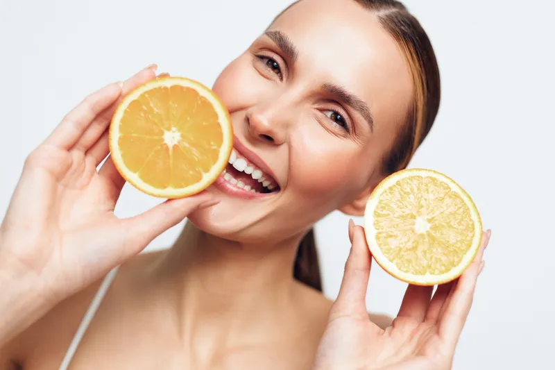 A smiling young woman holds two halves of a fresh orange close to her face, showcasing healthy skin and bright fruit. Concept for beauty, health, and natural wellness.