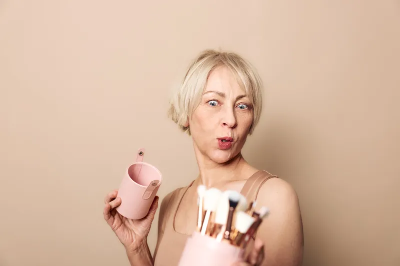 Mature woman with short blonde hair holds pink cup and makeup brushes, posing on beige background. Concept for beauty routine, skincare, self-care, and cosmetics for older women.