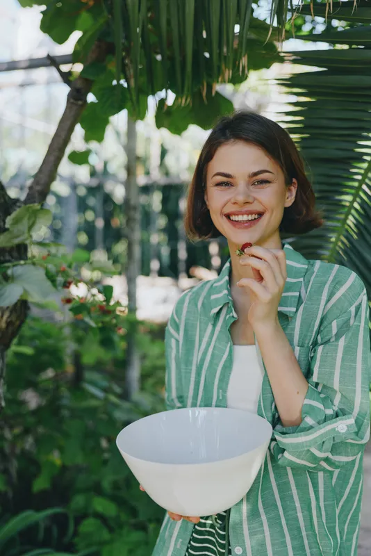 A cheerful woman holds a white bowl outdoors, smiling warmly as sunlight filters through lush foliage.