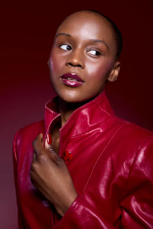 Portrait of an elegant black woman with dark skin wearing a red leather jacket, looking sideways with glossy lips and smooth skin. Studio lighting highlights her confident and stylish mood.