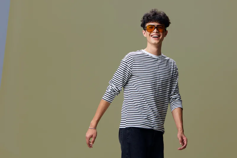Smiling young man wearing a striped shirt and orange sunglasses, standing in a relaxed pose with curly hair and a neutral background, casual style, joyful mood.