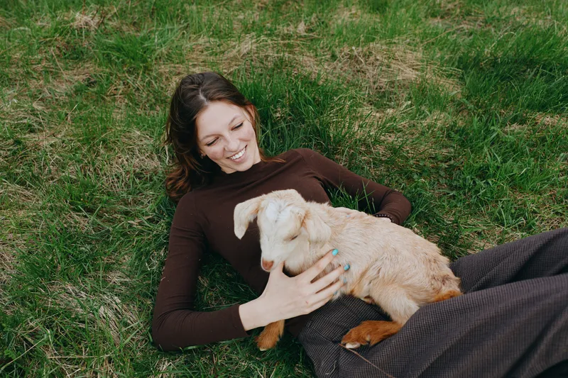 Happy woman lying on grass beside a playful goat kid, embracing nature and friendship in an outdoor lifestyle moment, warm sunlight, relaxed mood, gentle bonding between human and animal