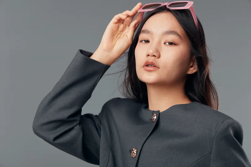 Young woman with long dark hair wearing a dark coat and pink sunglasses poses against gray background in studio. Closeup portrait with confident expression and stylish fashion accessory.