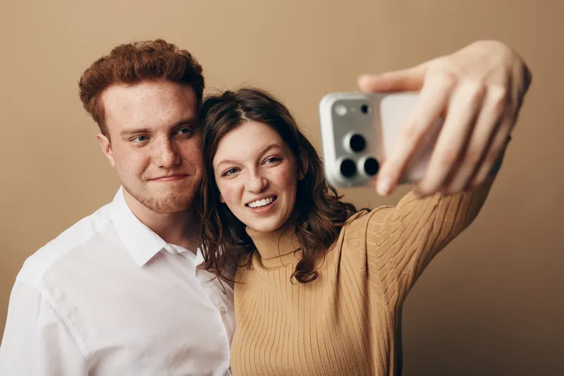 Young couple with red hair and brown hair takes selfie with smartphone on beige background. They smile and pose closely, capturing happy moment of friendship and modern lifestyle together.