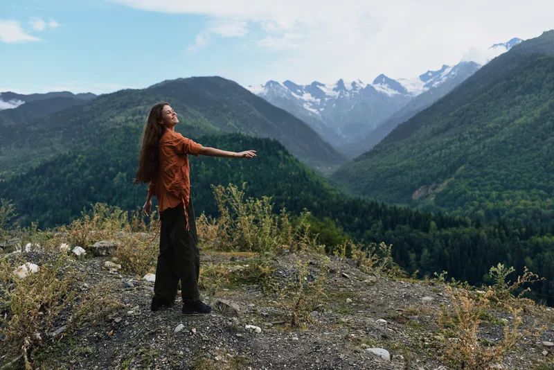 Woman standing on mountain ridge with outstretched arm looking at valley authenticity inclusivity long hair hiking clothes alpine landscape calm