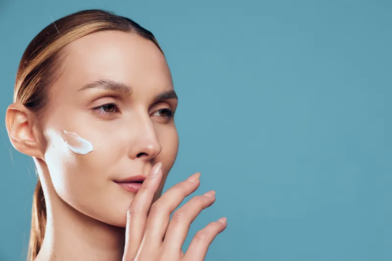 Skincare woman applying white face cream on cheek, closeup portrait, blue background, wellness concept, self care routine, soft studio light, clean glowing healthy skin