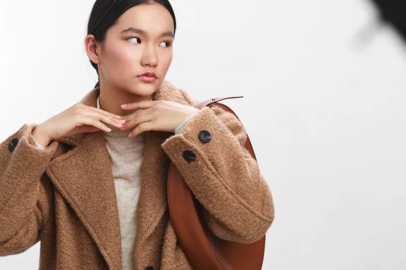asian woman wearing a warm brown coat in a studio, adjusting her collar with a thoughtful gaze, conveying fashion confidence and a calm, contemporary portrait mood.