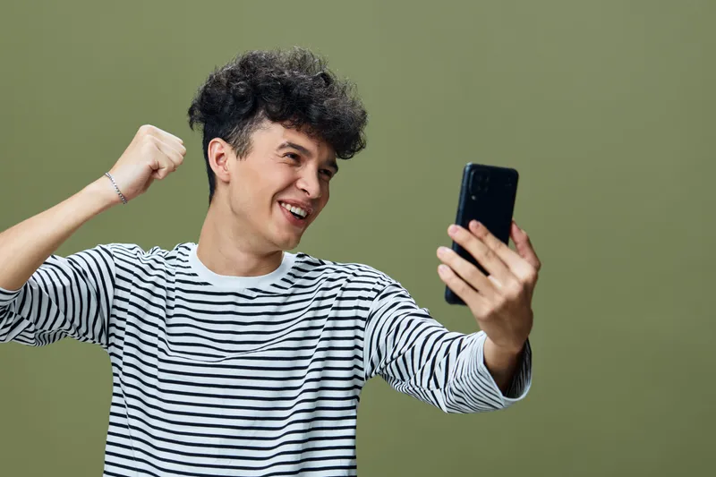 Young man smiling joyfully while looking at smartphone, wearing a striped shirt against a green background, expressing happiness and excitement.
