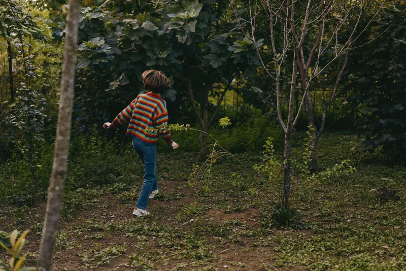 Boy child in a striped sweater walking through a green forest, age nine, joyful and curious, casual outfit with jeans and sneakers, exploring nature in a moody outdoor lifestyle scene.