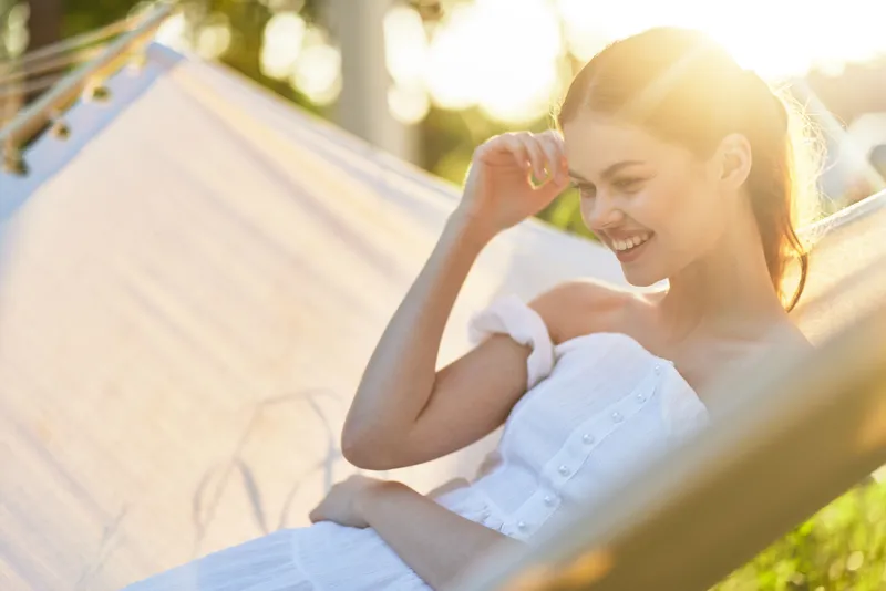 Young woman in white dress relaxing in sunlit hammock, enjoying serene summer moment, with a joyful expression and soft natural background