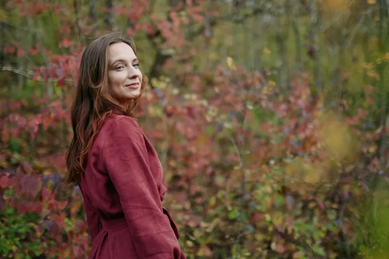 A young woman in a red coat smiles naturally among autumn foliage, a candid portrait emphasizing authenticity and realistic skin tones, fabric texture, soft depth and warm ambient light.