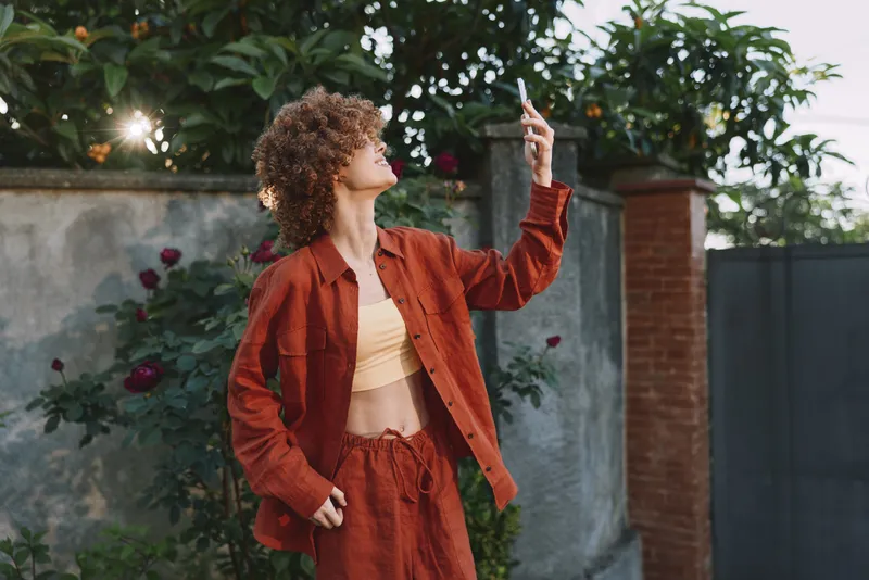 Young woman with a rust red jacket stands outdoors, looking up at her phone with a soft smile in a sunlit garden